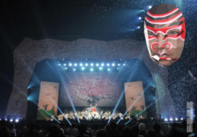 ステージ上の大きな顔型のスクリーンに隈取が重畳された顔を投影した写真