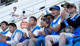Image: Photograph of volunteer athlete supporters comprising family members and co-workers cheering on the athletes. Wearing supporter uniforms, they are clapping and cheering as they watch the track and field action.