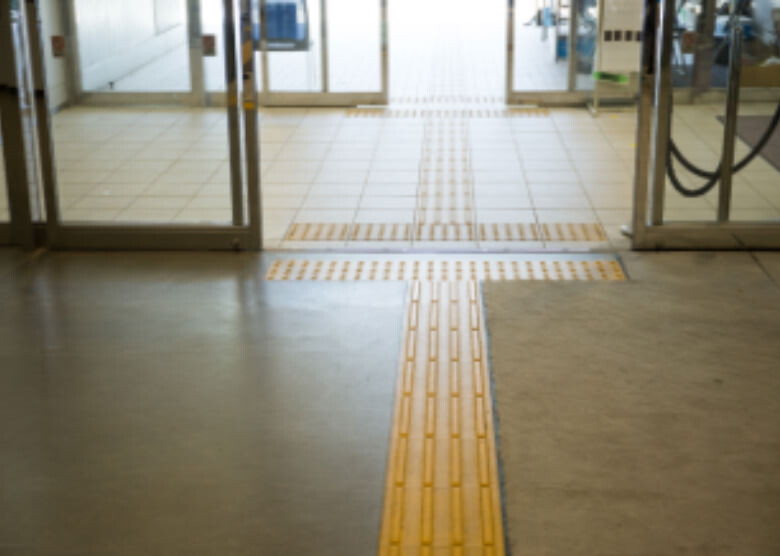 Image: Photograph of the entrance to the Machida Stadium in Nozuta Park, Machida City, Tokyo, showing continuous Braille blocks leading from in front of the automatic doors into the stadium.