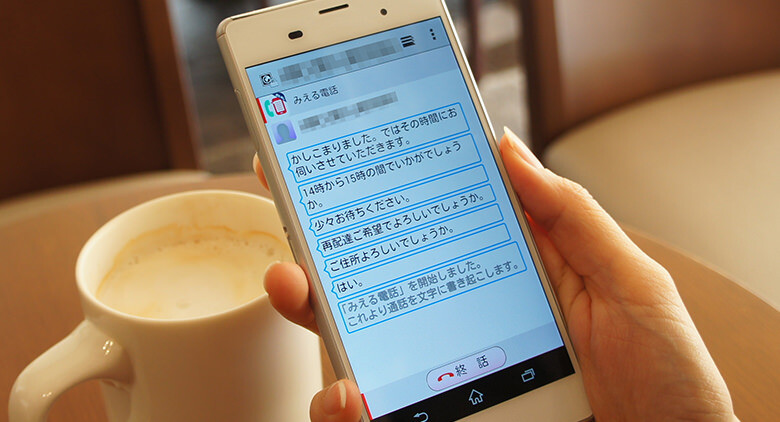 Image: Photograph of a hand operating Docomo's Mierudenwa. The app converts the voice of the person the user is speaking to into text and displays their words on the talk screen.