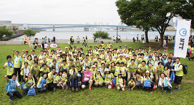 Image: NTT staff and several tens of people gathered together on the river terrace for a group photograph.