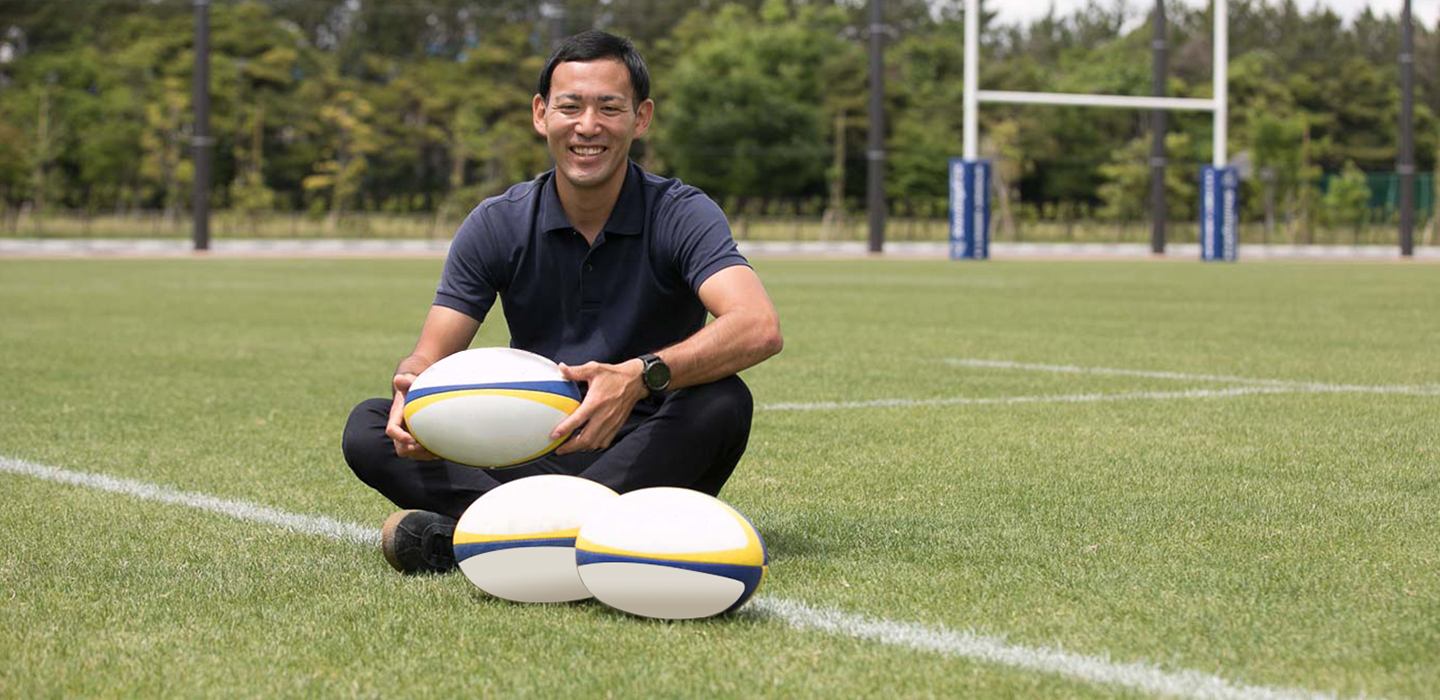 Image: A smiling Mr. KAWAHARA holds a football as he sits on a football field