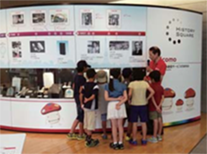 写真：NTTドコモ歴史展示スクエアで、スタッフが説明し、子どもたちが見学する様子