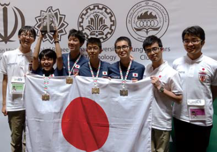 写真：情報オリンピックのパネルの前でメダルと日本国旗を掲げる生徒たち