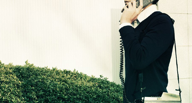 画像：ショルダーフォンをかける男性の写真。モバイル通信が誕生する前の1980年代は、外で通話するには大きなショルダーフォンを肩にかけなければなりませんでした。