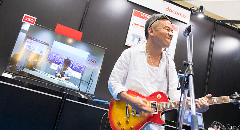 Image: Photograph of a man enjoying an online jam session in real time using 5G technology. The man is playing guitar and enjoying a jam session with another man playing keyboard in a distant location whose image is shown the monitor. 