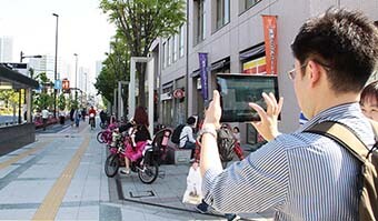 画像：街中でタブレットで写真を撮り記録するバリアフリー調査の男性の後ろ姿の写真。